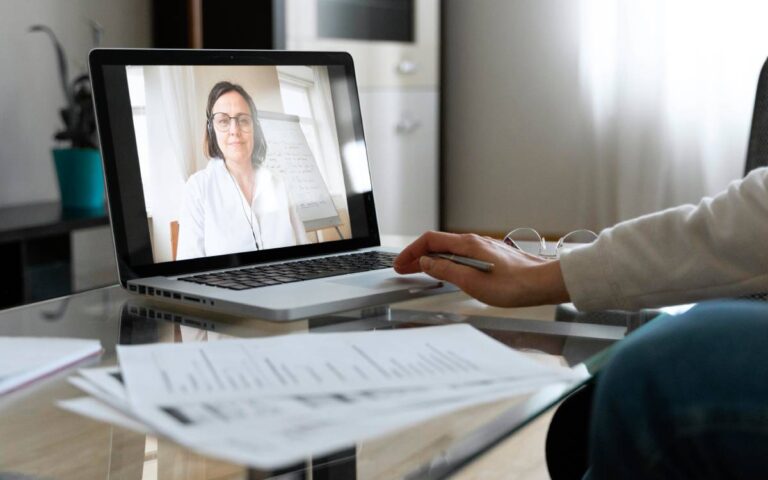 Pessoa assistindo uma aula online em um laptop com gráficos e documentos sobre a mesa.