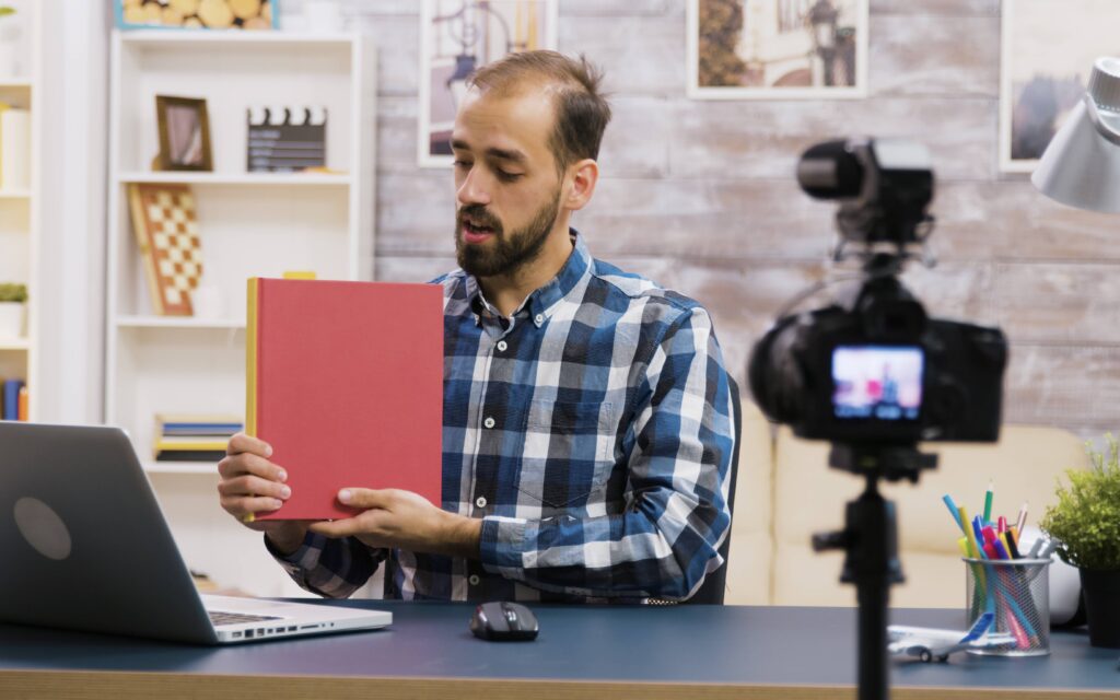 Homem gravando uma aula online com um livro em mãos, utilizando um laptop e uma câmera.