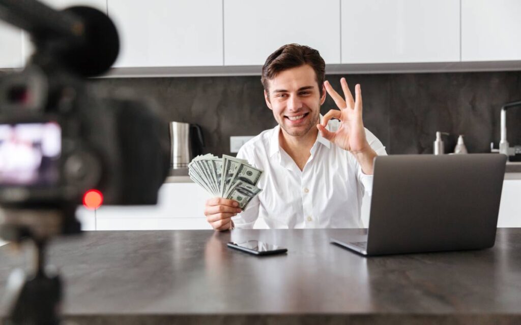 Homem segurando dinheiro fazendo sinal de ok em frente a uma câmera e um laptop, simbolizando monetização no TikTok.