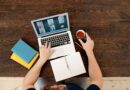Uma pessoa participando de uma reunião virtual em um laptop com café e cadernos em uma mesa de madeira.