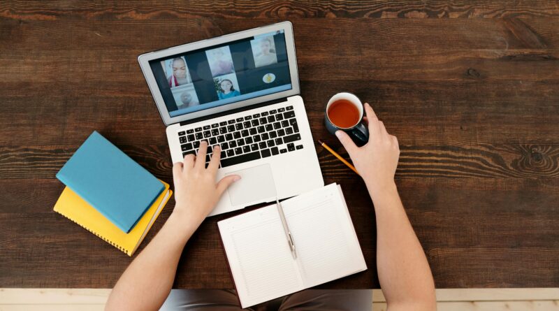 Uma pessoa participando de uma reunião virtual em um laptop com café e cadernos em uma mesa de madeira.