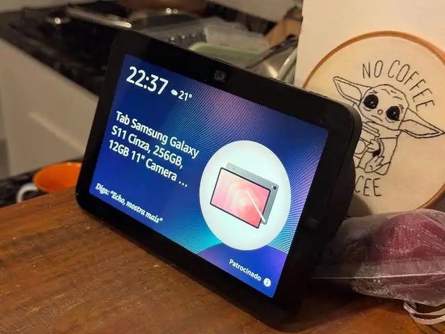 Detalhe do Echo Show com banner publicitário em destaque na tela ambiente, sobrepondo fotos e widgets